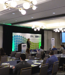 Conference room presentation: audience seated in rows facing a stage with a podium and a green/blue patterned backdrop.